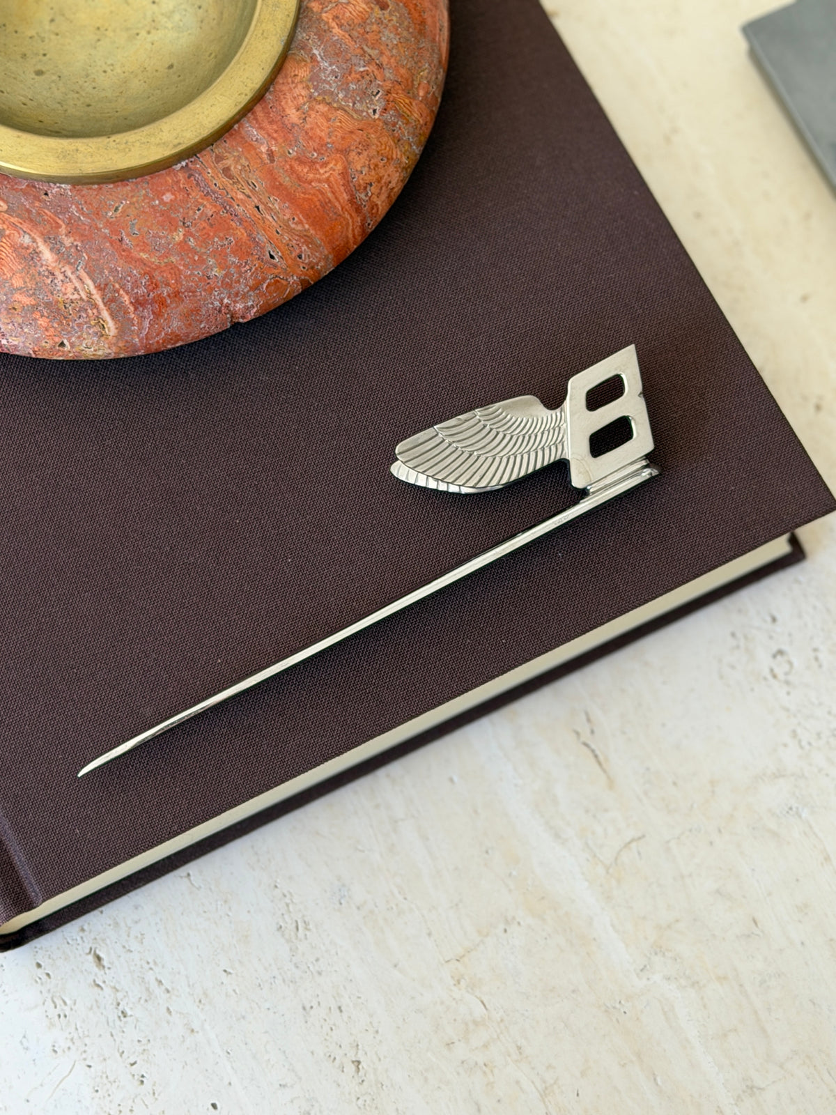 Overhead view of the Bentley letter opener on a brown clothbound book beneath a red stone ashtray, the feathered wing catching the light