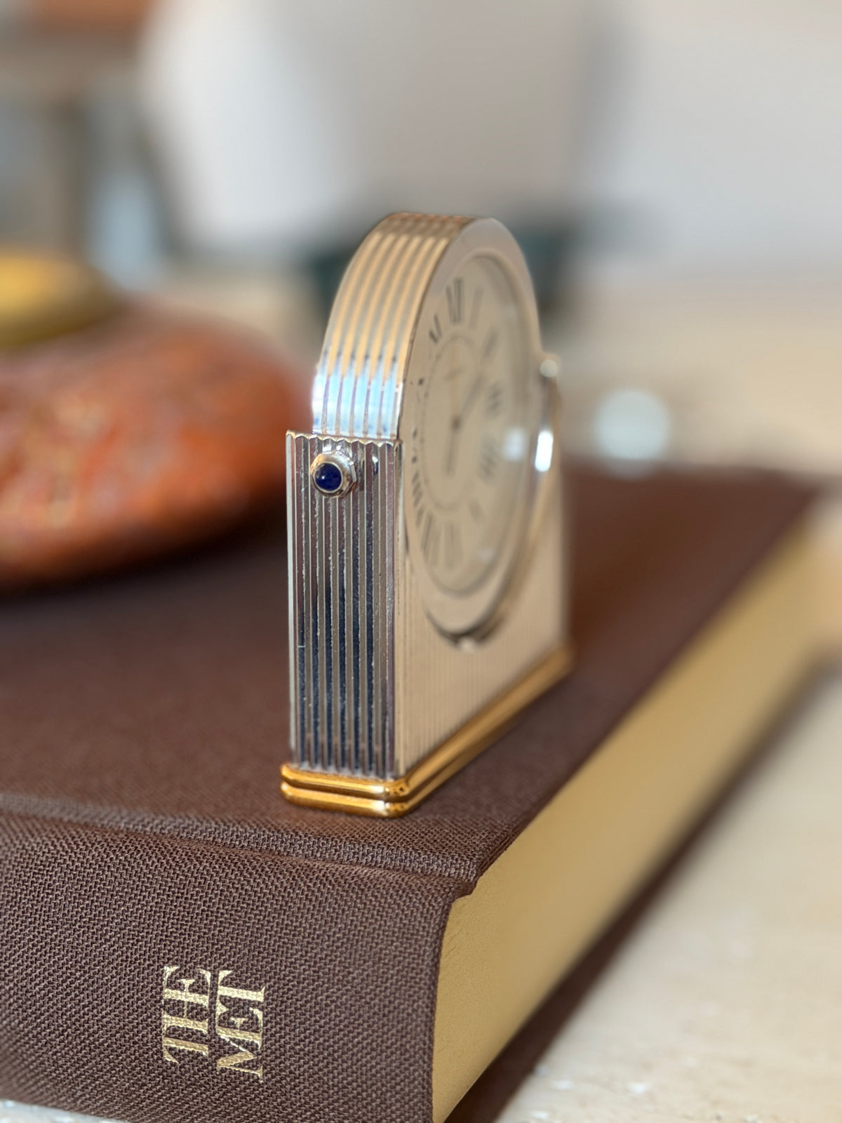 Side profile of the Cartier clock highlighting the ribbed edge of the round dial housing, a blue sapphire cabochon, and the gold-tone base