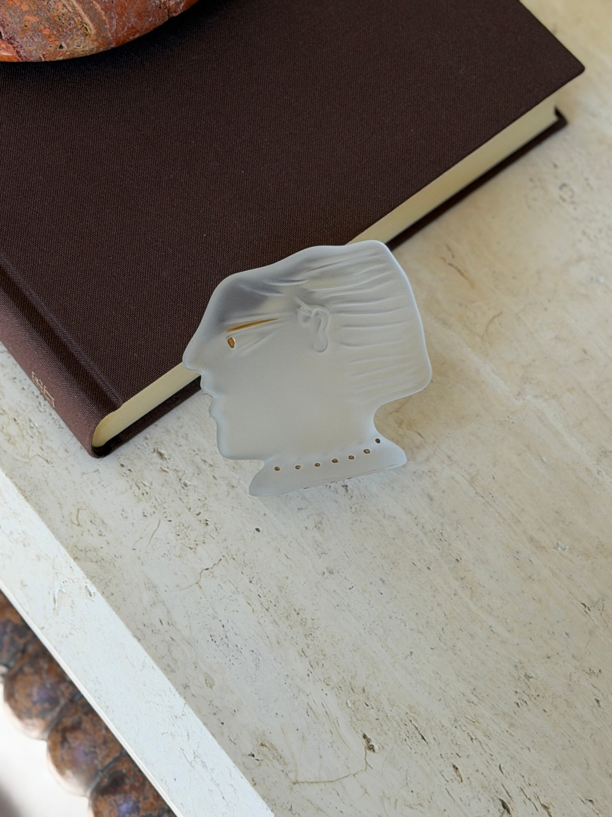 Daum frosted crystal head by Alekos Fassianos in profile, with a gold accented eye and dotted gold collar, leaning against a brown book on travertine