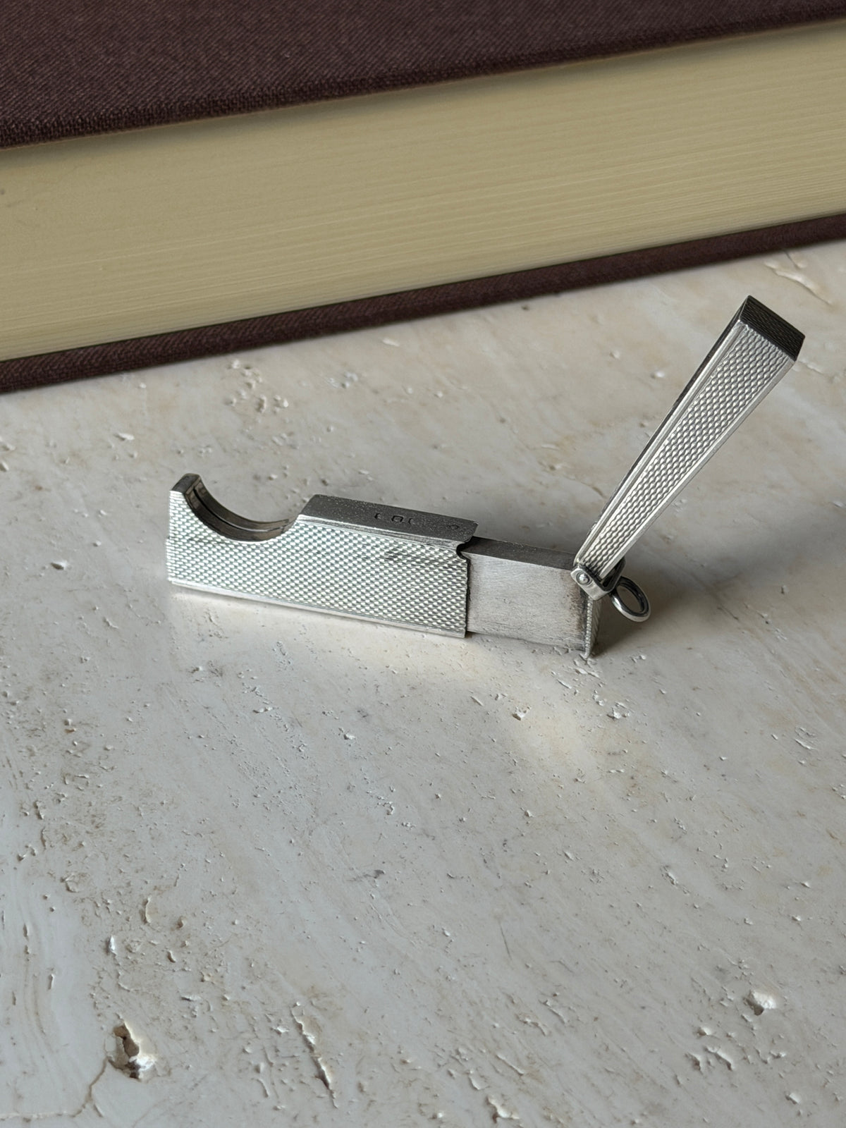 Dunhill cigar cutter opened on travertine, the engine turned lid raised and the curved guillotine slot exposed beside the sliding blade
