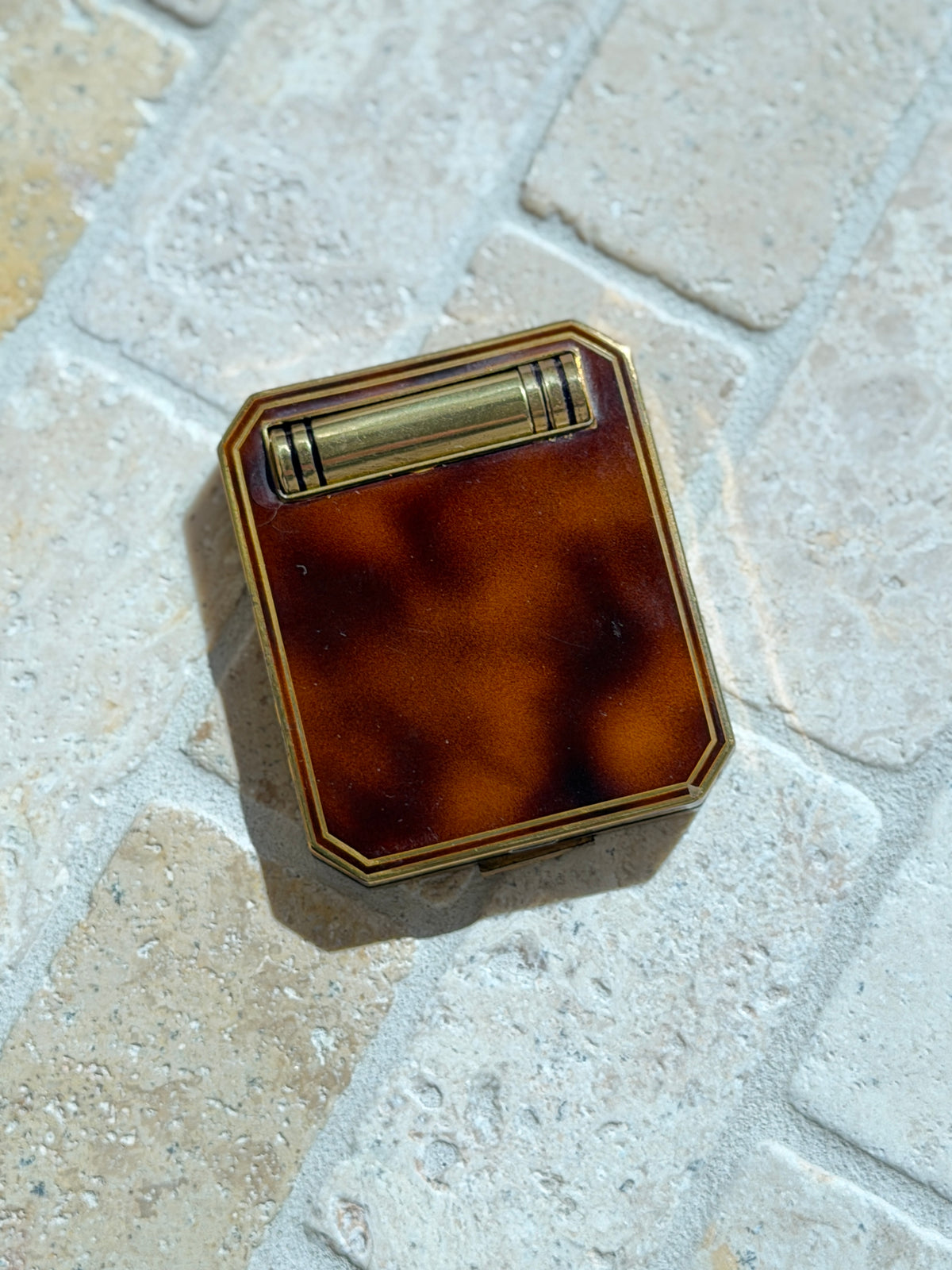 French Art Deco compact with a mottled tortoiseshell lid framed in brass, a banded brass lipstick tube set across the top, on tumbled stone pavers