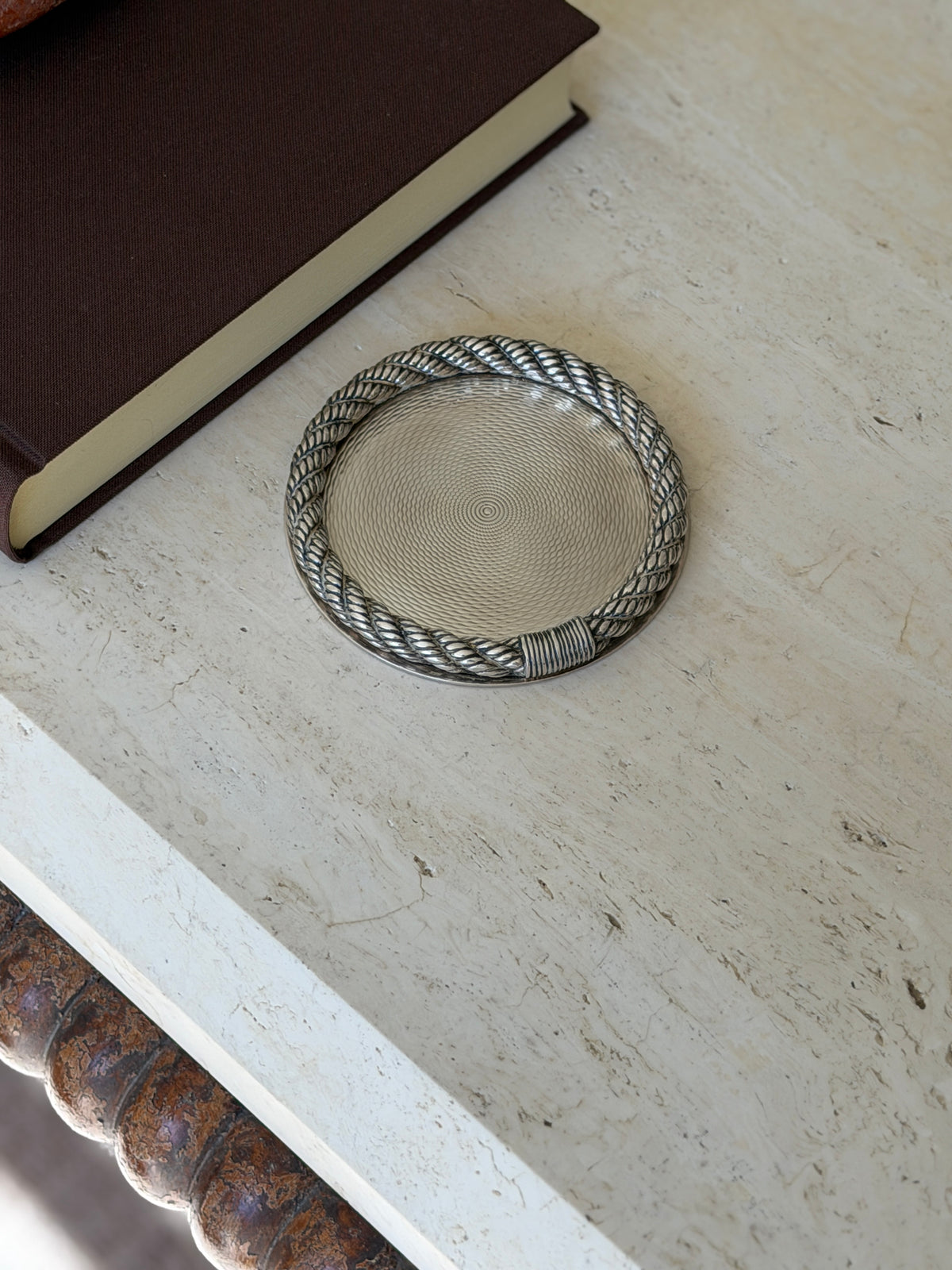 Hermes rope rim coaster on a travertine console near a brown book, the bobbin turned table leg visible below