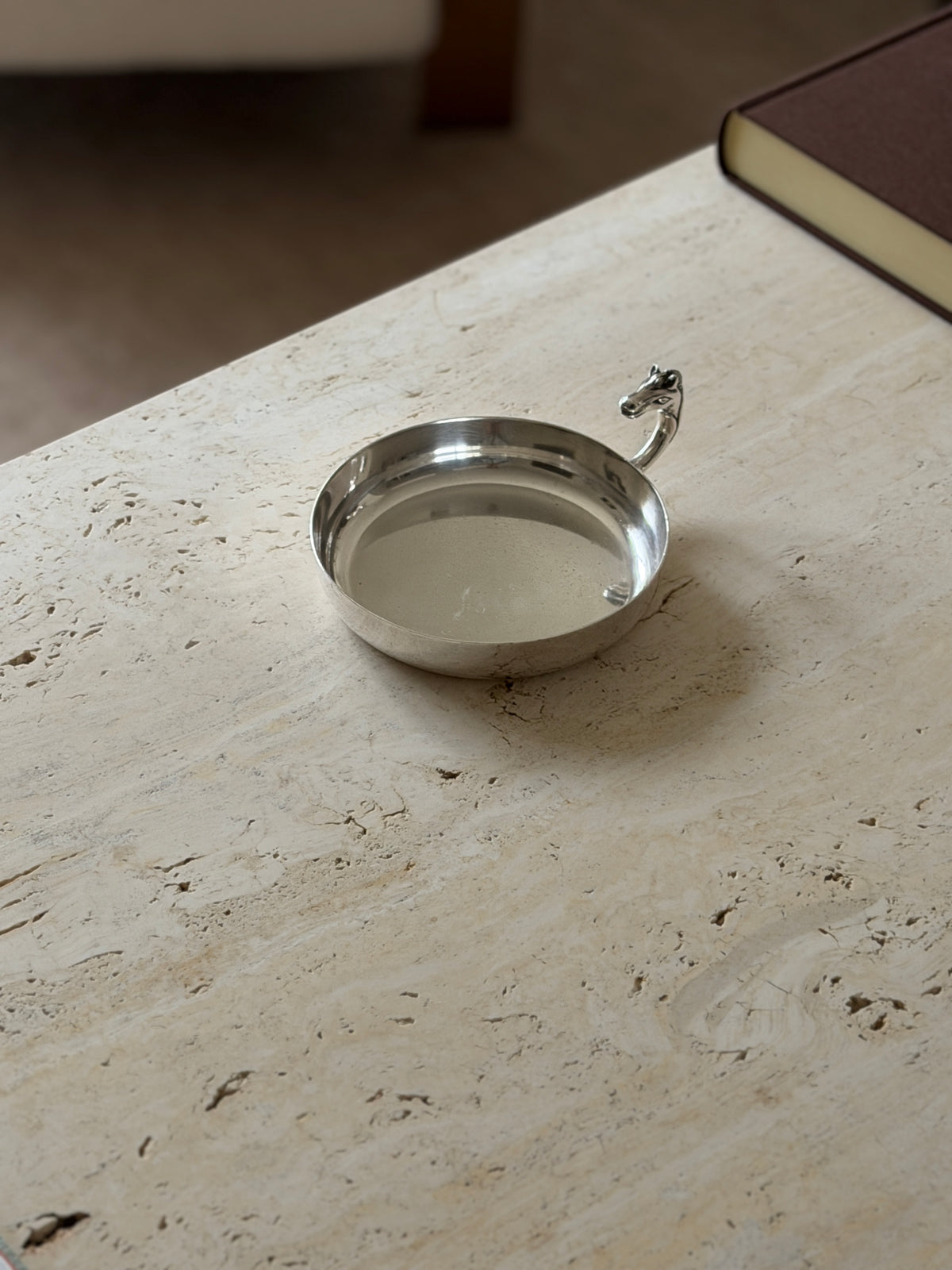Overhead view of the round silver dish on travertine, its polished interior and small horse head handle with a brown book in the corner