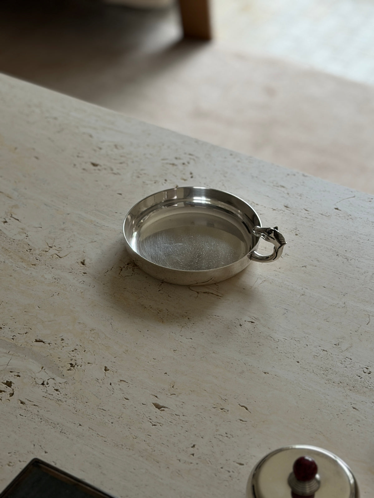 Silver catchall viewed from the opposite side, the horse head handle curling over the rim with a lidded silver box in the foreground