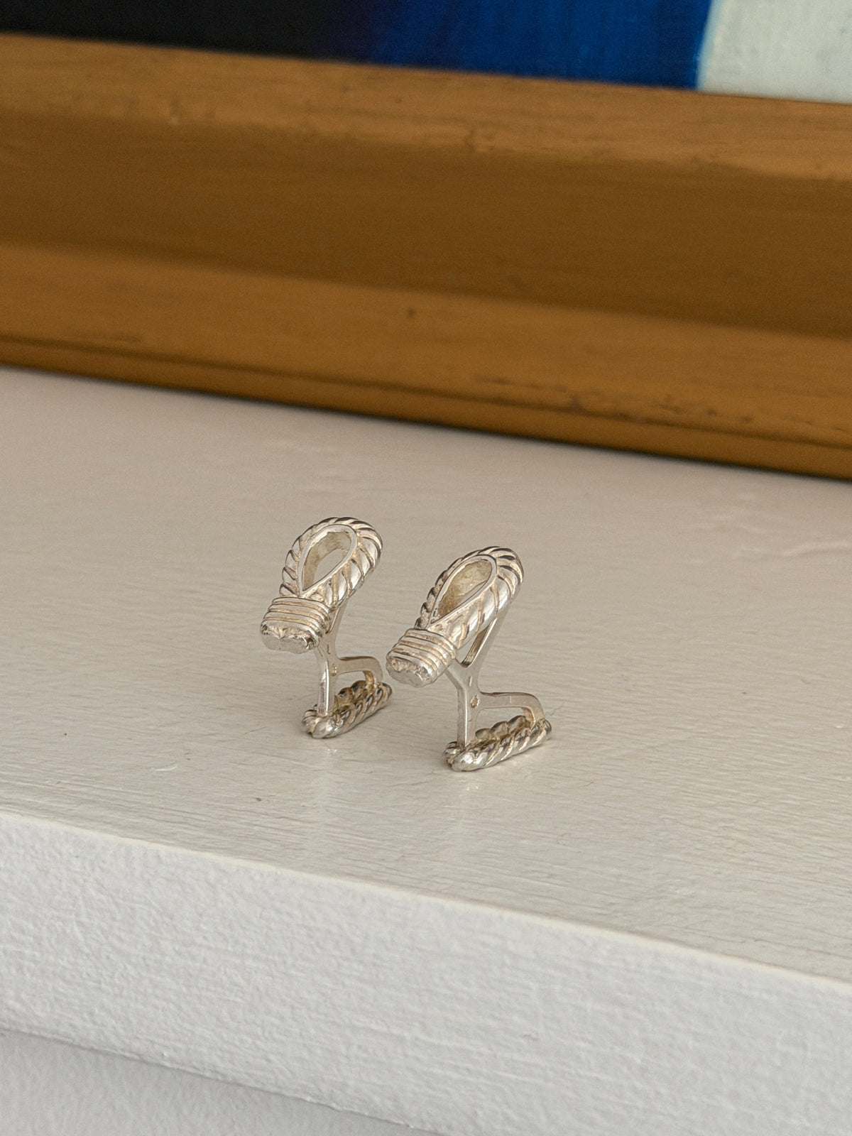 Pair of Hermes silver rope loop cufflinks standing on a white painted ledge, showing the wrapped collars and twisted rope backs