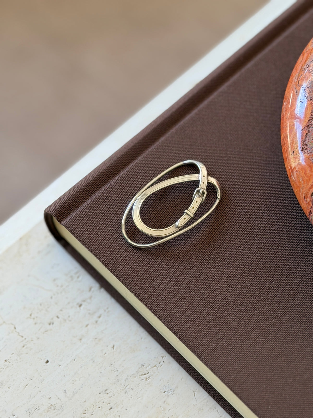 Hermes silver money clip laid diagonally on a brown linen book, the miniature buckle and pin holes catching the light