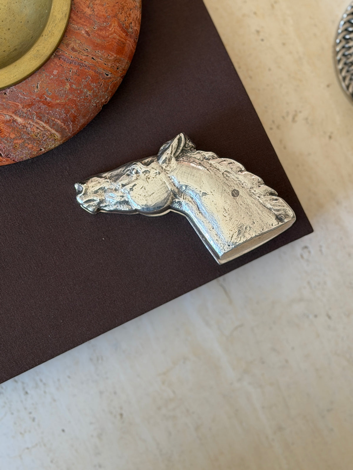 Sterling silver Hermes horse head paper clip resting flat on a dark book cover, shown in side profile with mane and ear detail visible