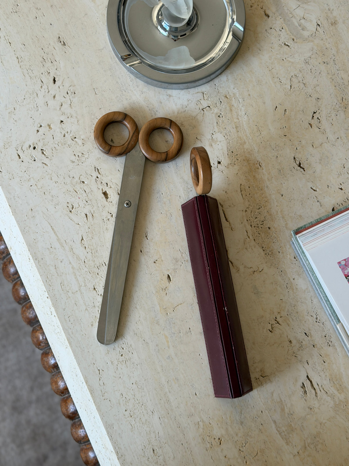 Overhead view of the Italian mid-century desk set, wood-handled scissors beside the letter opener sheathed in its oxblood leather case on a travertine surface