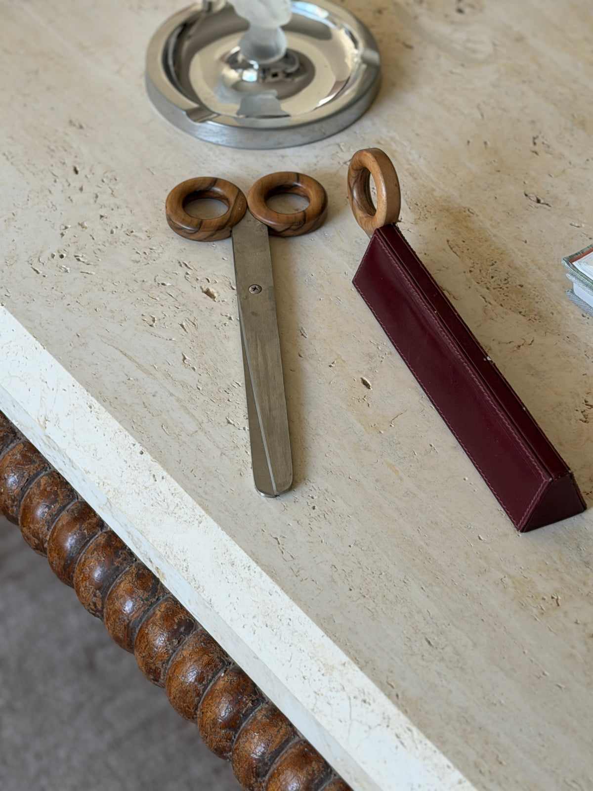 Angled view of the wood ring handled scissors and leather-cased letter opener laid side by side on travertine, with a chrome ashtray in the background