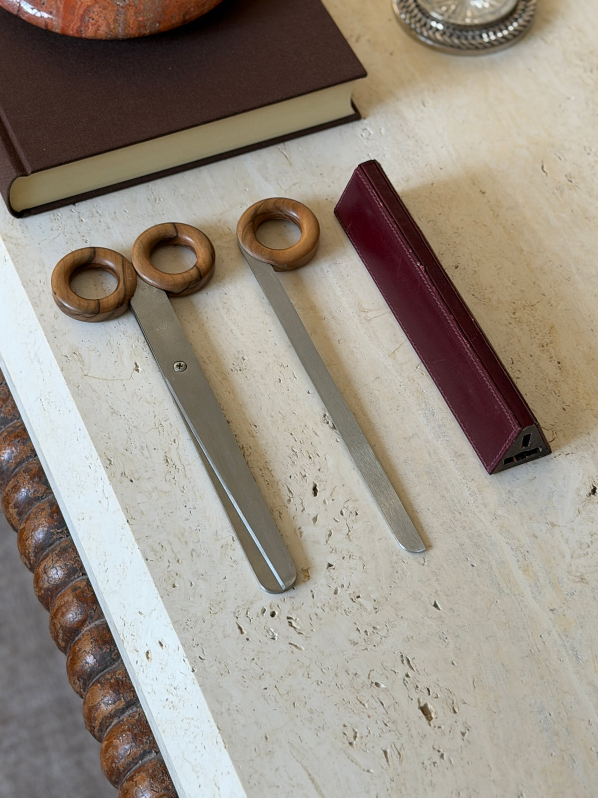 Complete desk set of scissors, letter opener, and triangular oxblood leather case with stitched edges, shown beside a brown book on travertine
