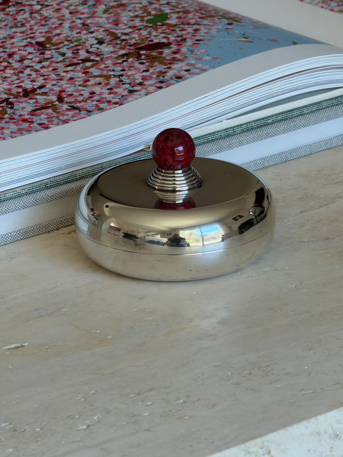 Front view of the Puiforcat candy dish on travertine, its mirror polished lid reflecting the room below the red finial