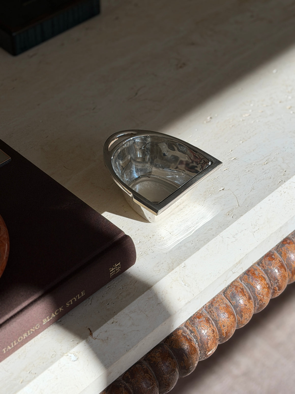 Ralph Lauren silver nut bowl shaped as a stirrup with a slotted top bar, on a travertine console in raking sunlight beside a brown book