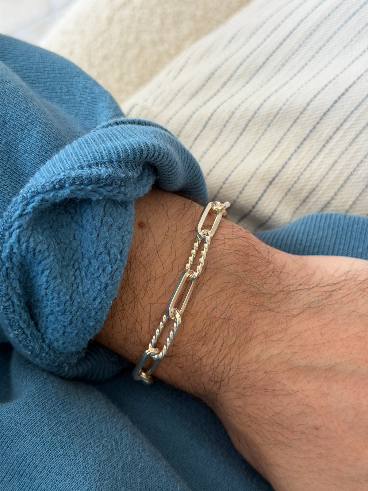 Bracelet worn on the wrist near a wooden table edge, cream ribbed top and blue sweatshirt cuff visible