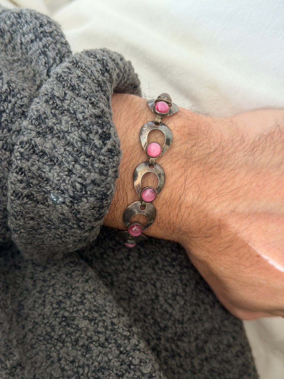 Pink cabochon bracelet worn over a gray sweater cuff, the hinged links and darkened patina along each curved panel