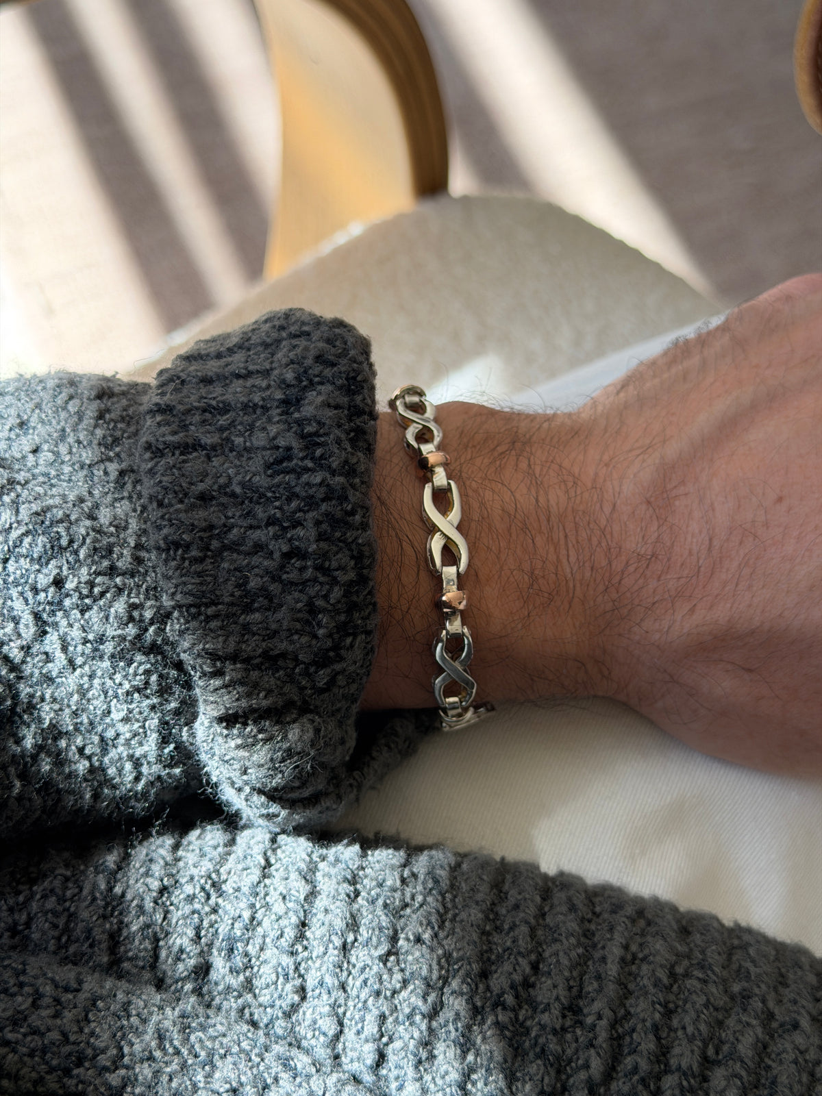 Two tone silver and gold bracelet on the wrist beside a bentwood chair, the infinity links reflecting the window light