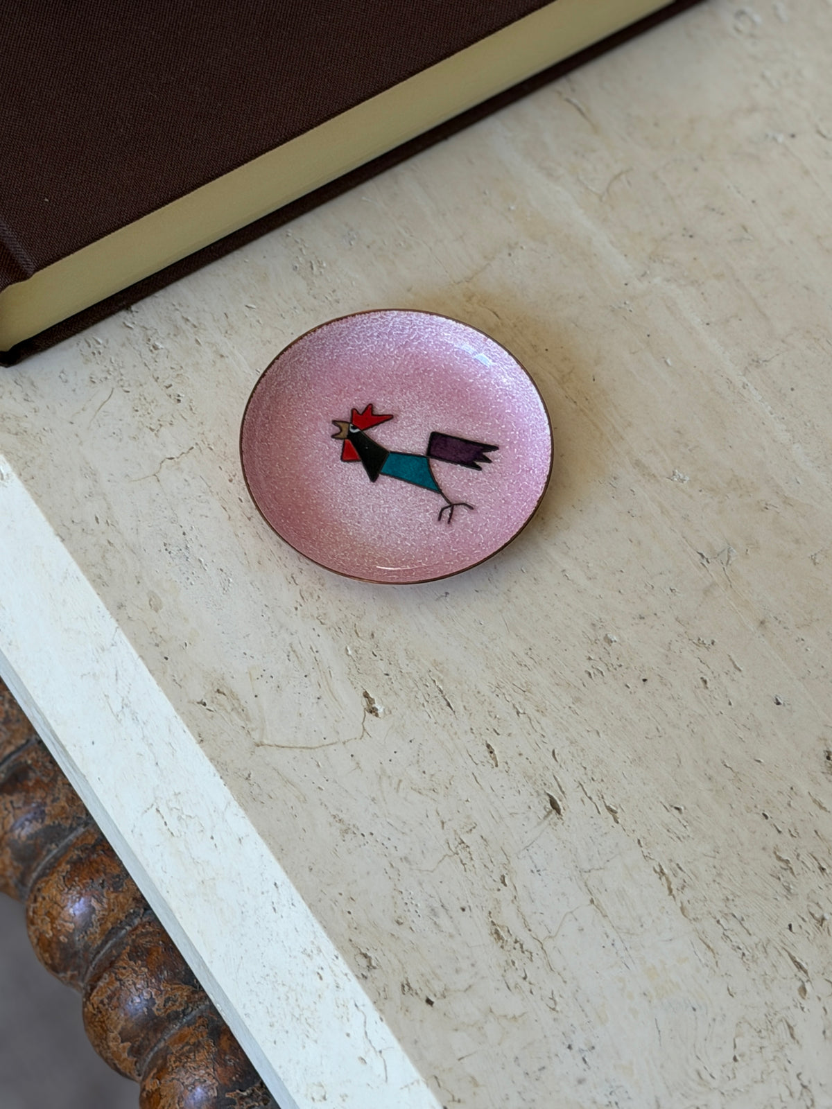 Overhead view of the small pink enamel dish with an abstract rooster design, on a travertine table beside a brown book