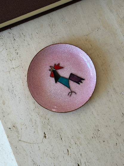 Overhead view of the enamel rooster dish, centered on the travertine table near the book edge