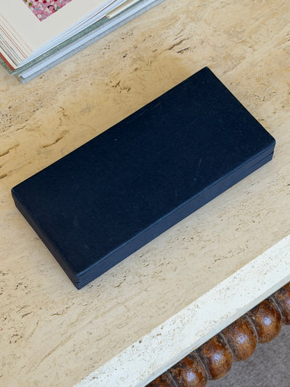 Closed navy textured presentation box on a travertine table, with a floral book visible behind it