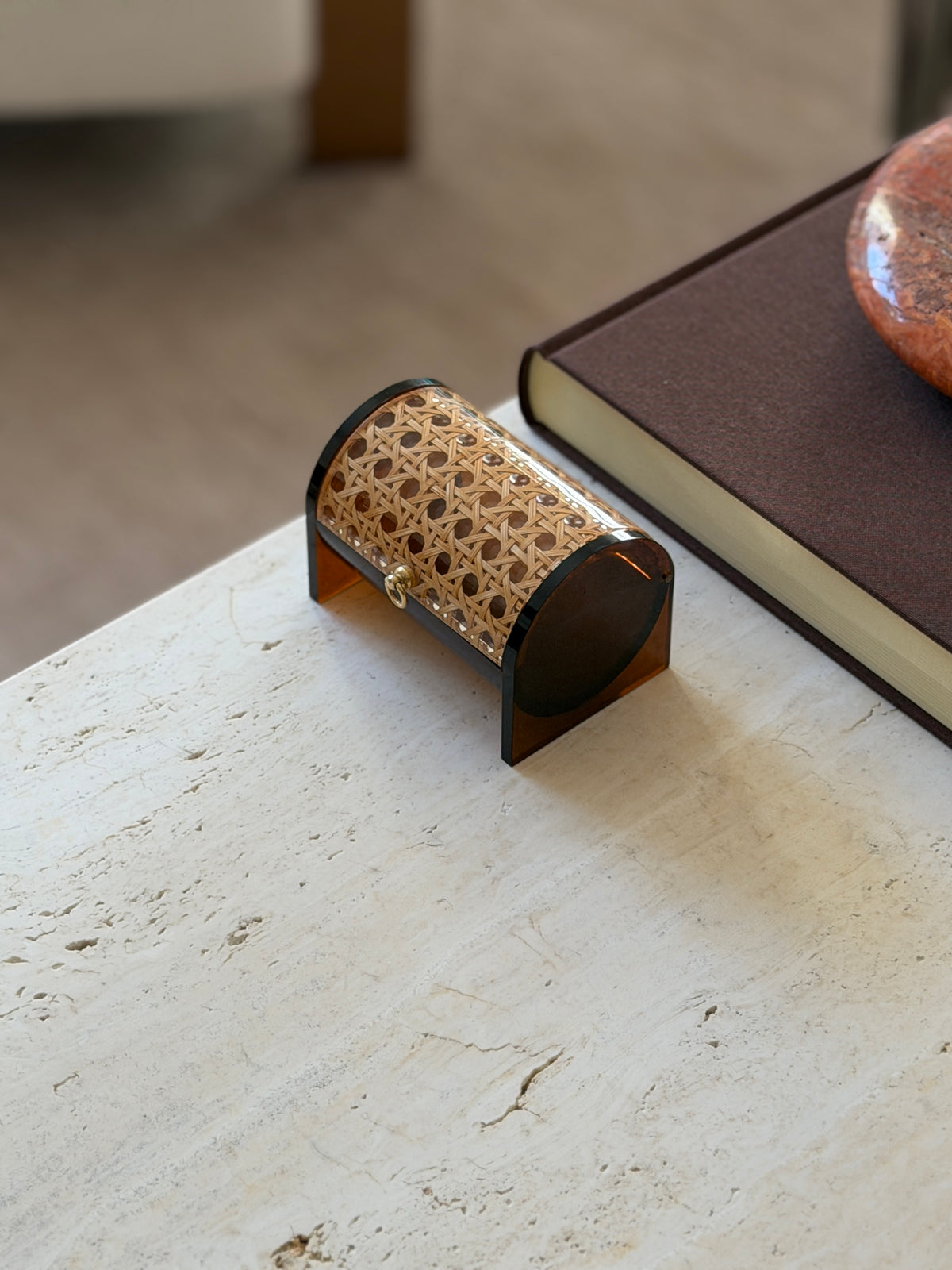 Closed cylindrical box seen from one end, the rounded amber Lucite end panel framing the cane-covered lid, a red stone vessel on a book behind