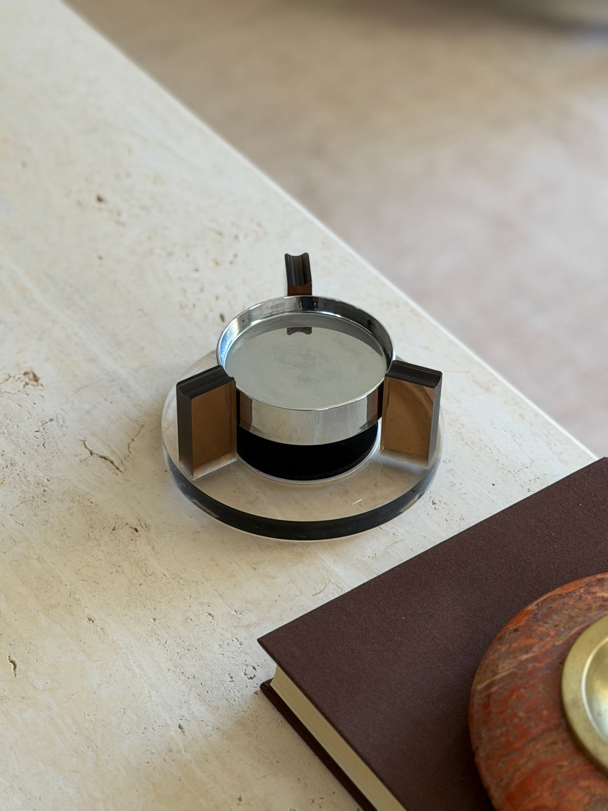 Round base magnetic ashtray at the corner of a travertine table, the dark gap between the chrome bowl and black base showing its floating effect