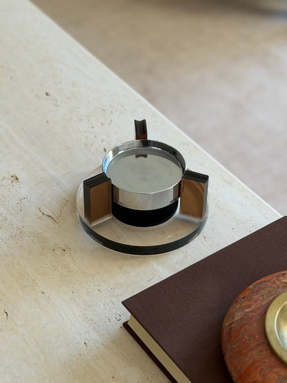 Round base magnetic ashtray at the corner of a travertine table, the dark gap between the chrome bowl and black base showing its floating effect
