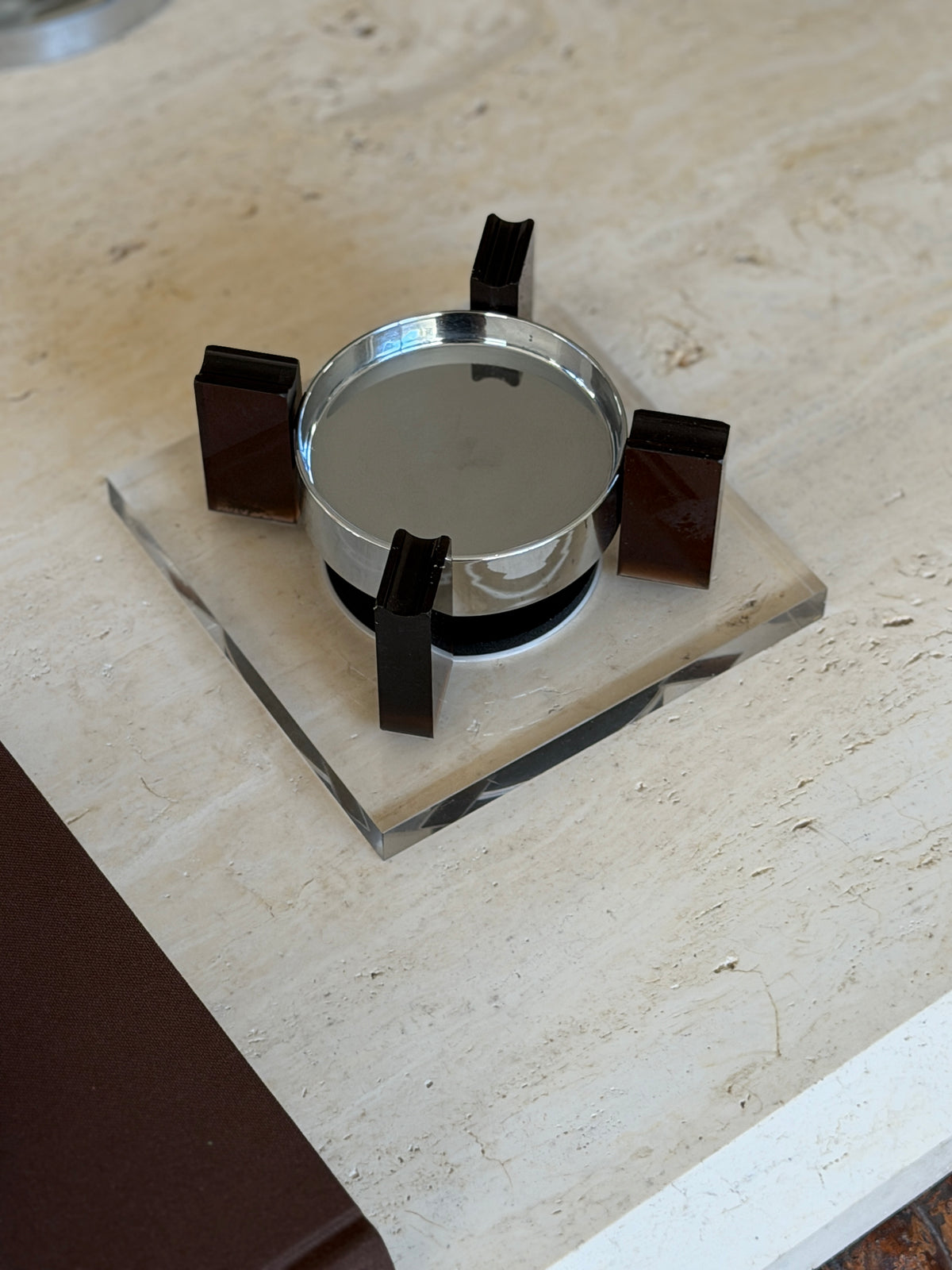 Top-down view of the square base ashtray set diagonally on travertine, the four amber uprights evenly spaced around the reflective chrome bowl