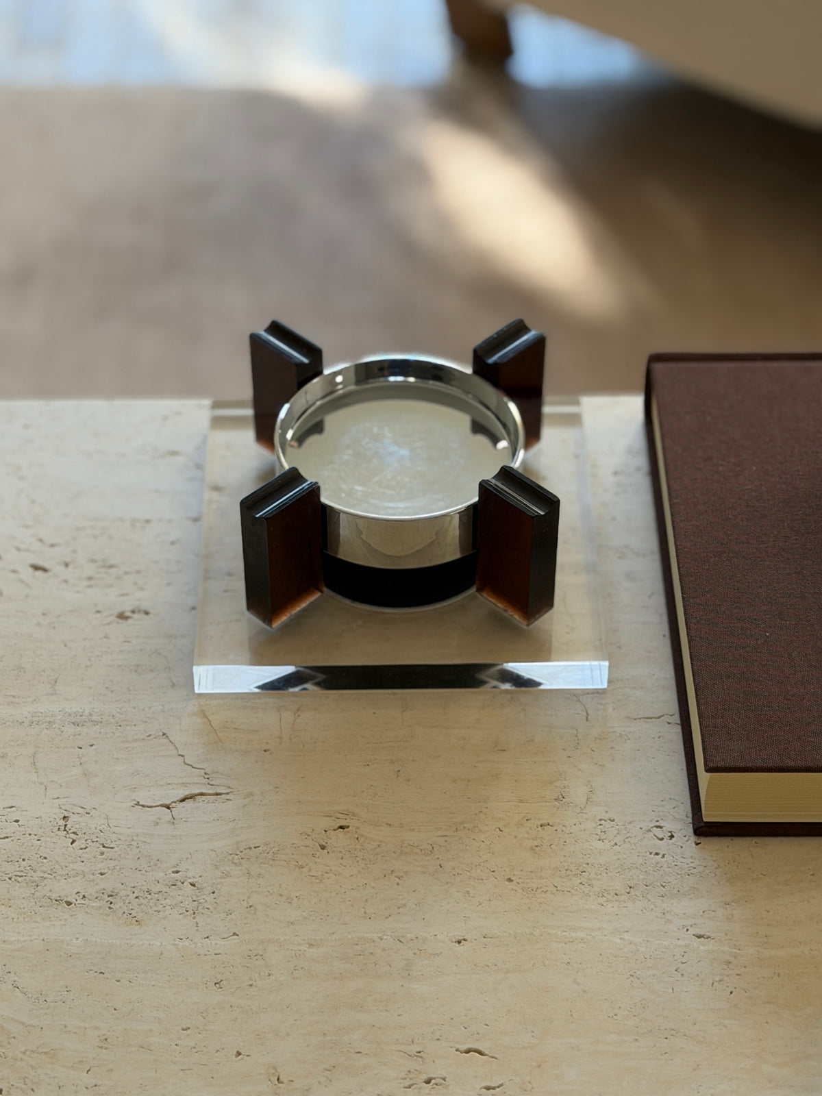 Square base magnetic floating ashtray seen at table level beside a brown clothbound book, light catching the clear Lucite edges