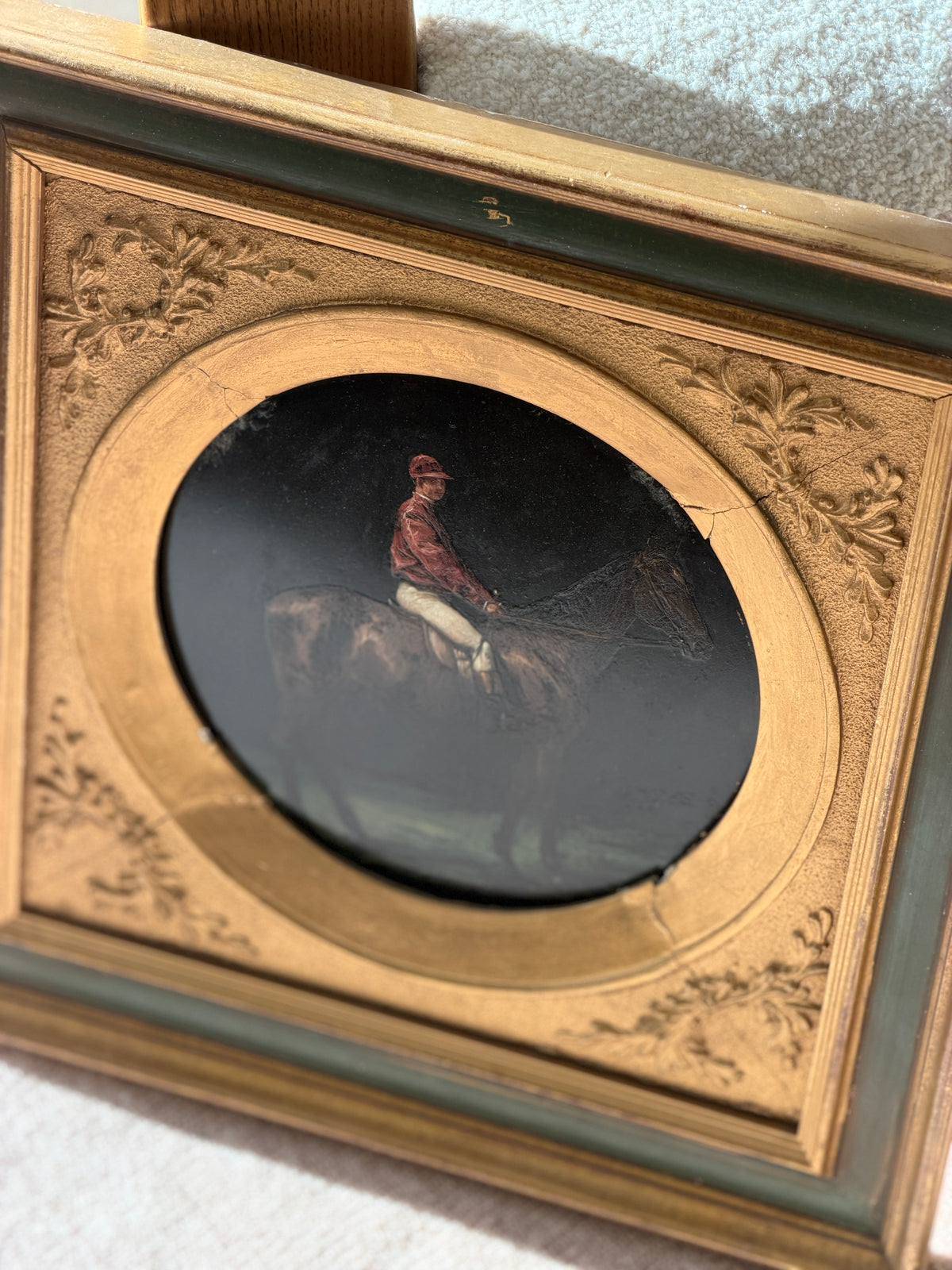 Antique oil painting of a jockey in a red silk jacket and cap on a bay horse, painted on a round dark plate within a gilt and green painted frame with relief foliage corners