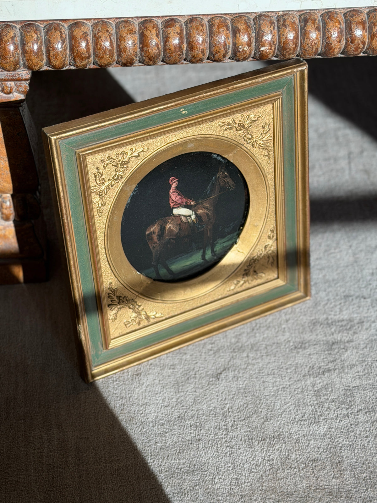 Angled view of the round jockey portrait under a carved wood table, showing the embossed oak leaf corners and patinated gilding