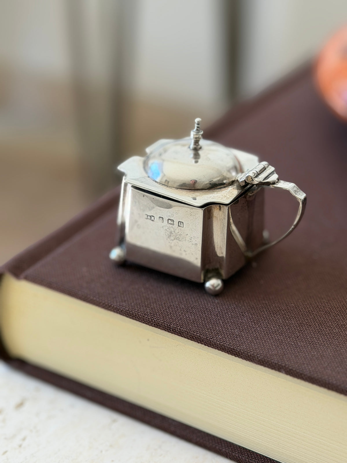 Antique sterling silver mustard pot with a domed hinged lid and baluster finial, a row of English hallmarks on the side, standing on ball feet on a brown book