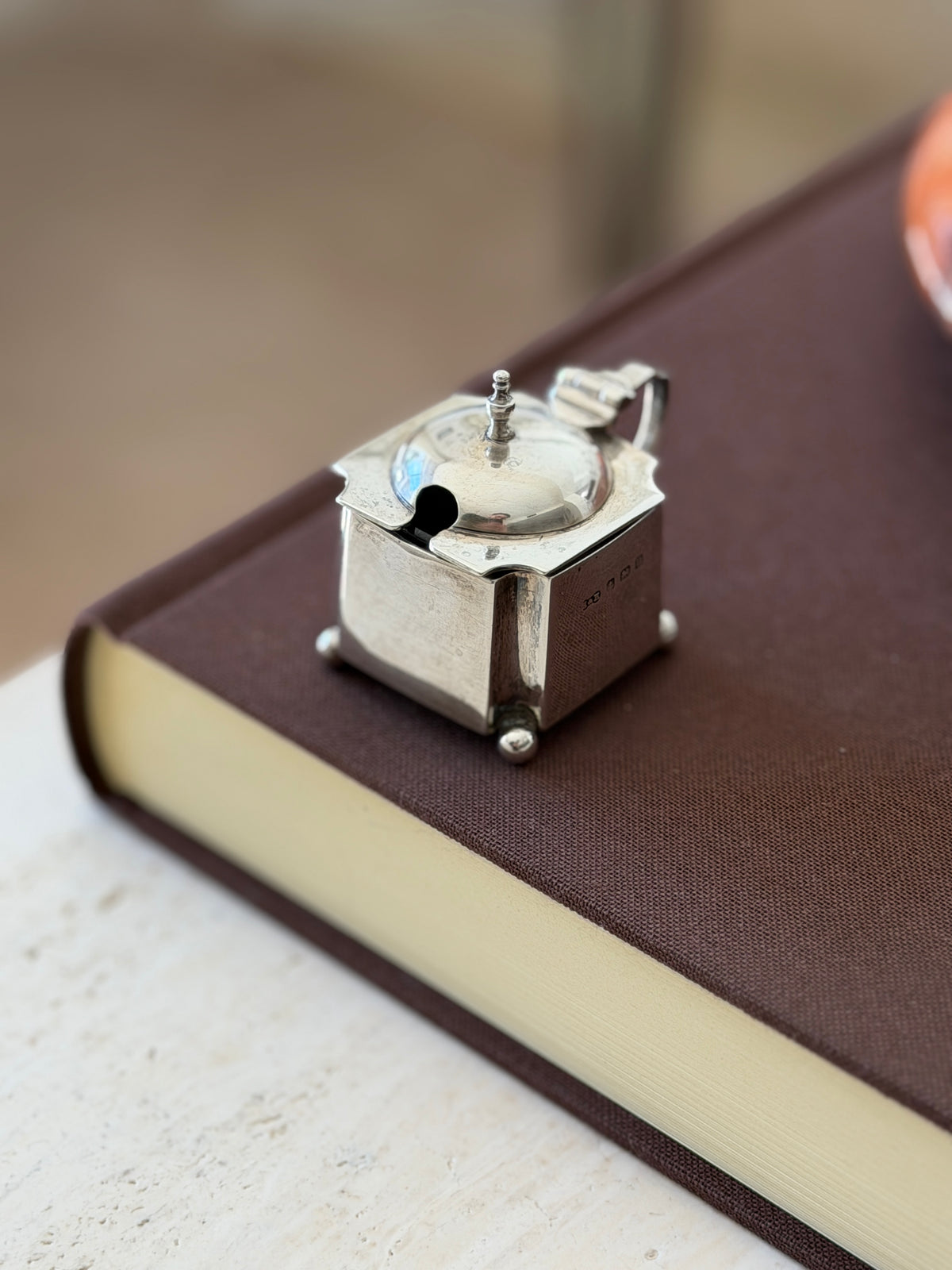 Angled view of the mustard pot on a book, the spoon slot and finial in front and a full set of hallmarks on the side panel