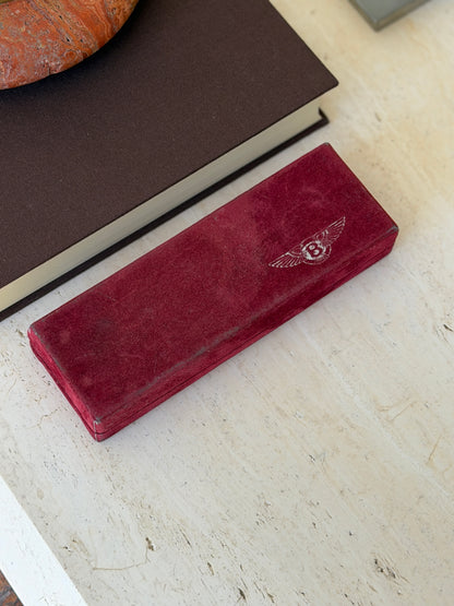 Burgundy suede presentation box for the Bentley letter opener, embossed with the winged B emblem, on a travertine table beside a brown book
