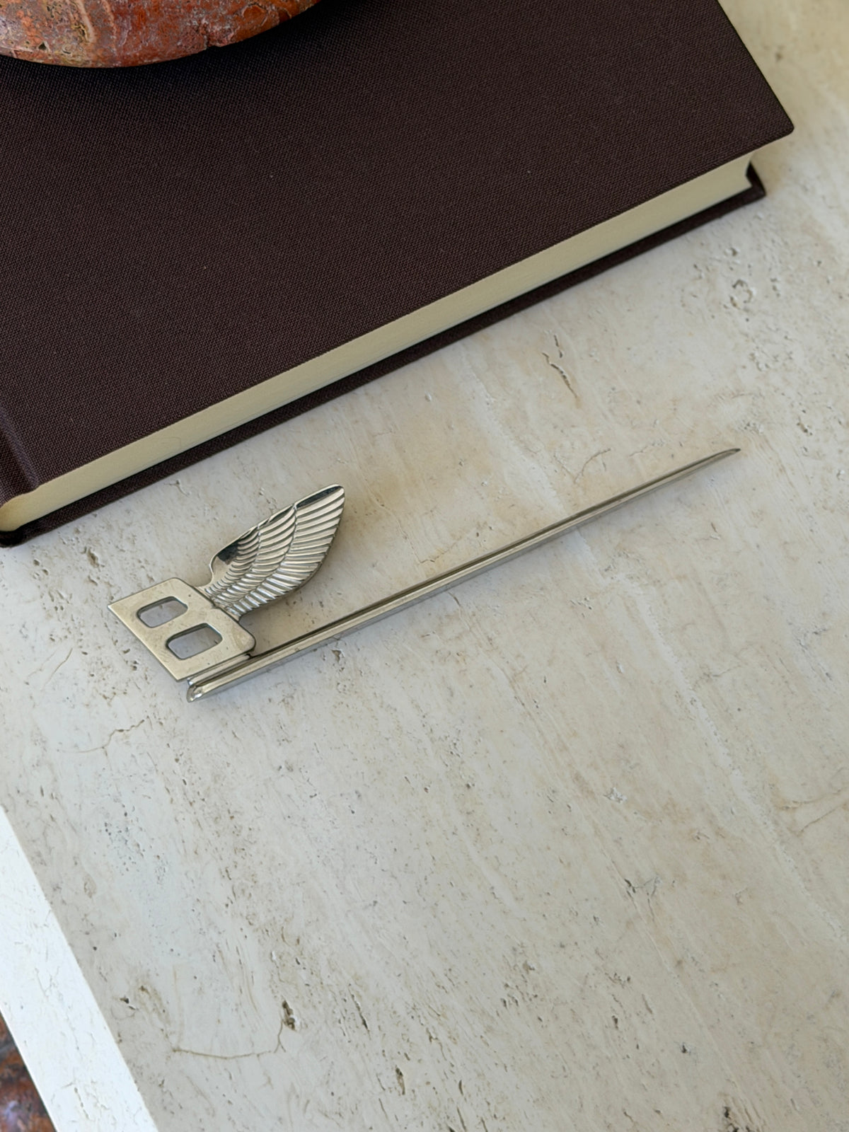 Bentley Flying B letter opener in polished silver plate, the winged B handle above a long slim blade, lying on travertine
