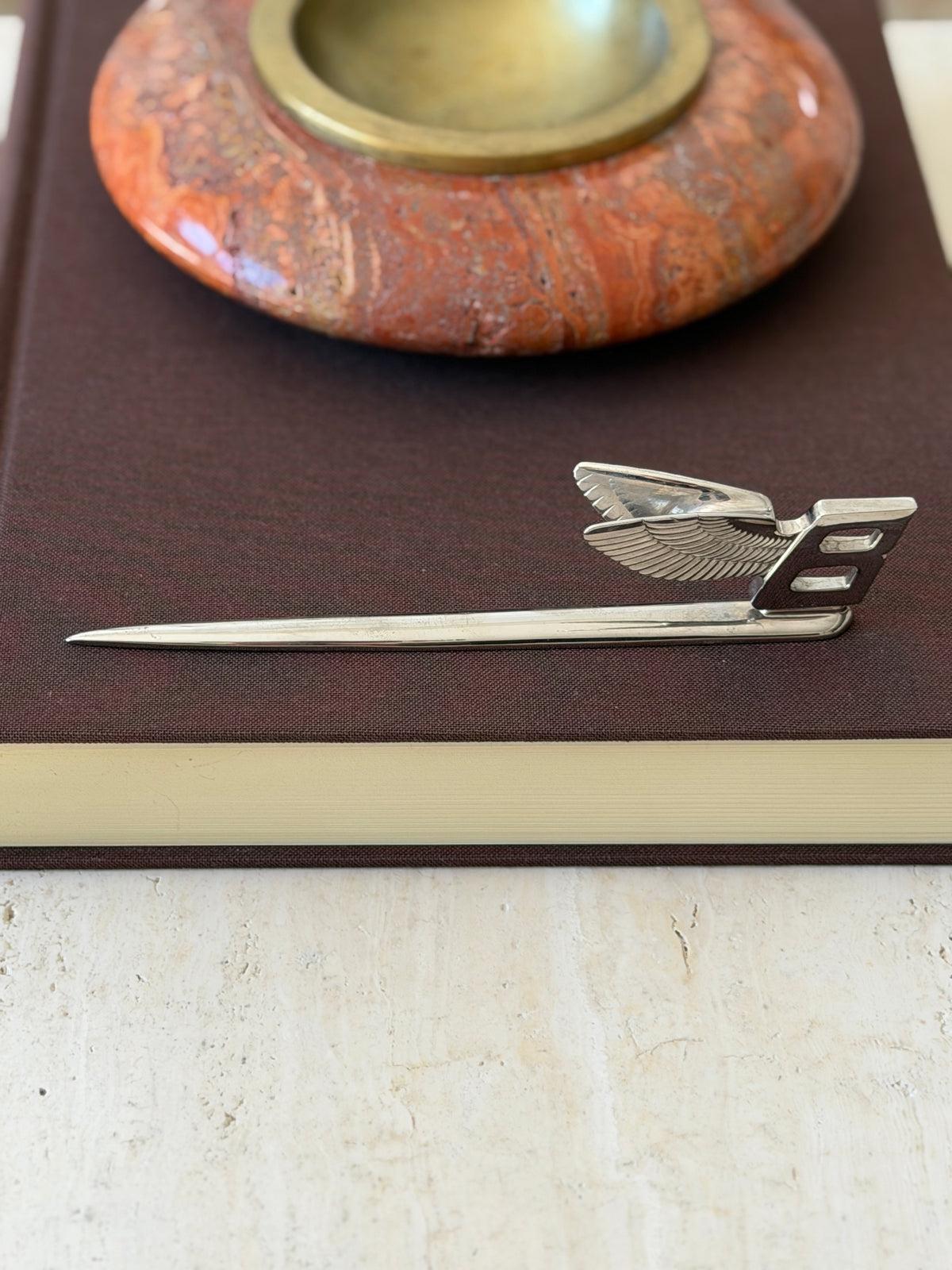 Side profile of the Flying B letter opener on a brown book, the swept wing rising above the tapered silver plate blade