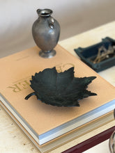 Angled desk display view of dark oxidized bronze leaf tray styled on a book stack alongside decor objects