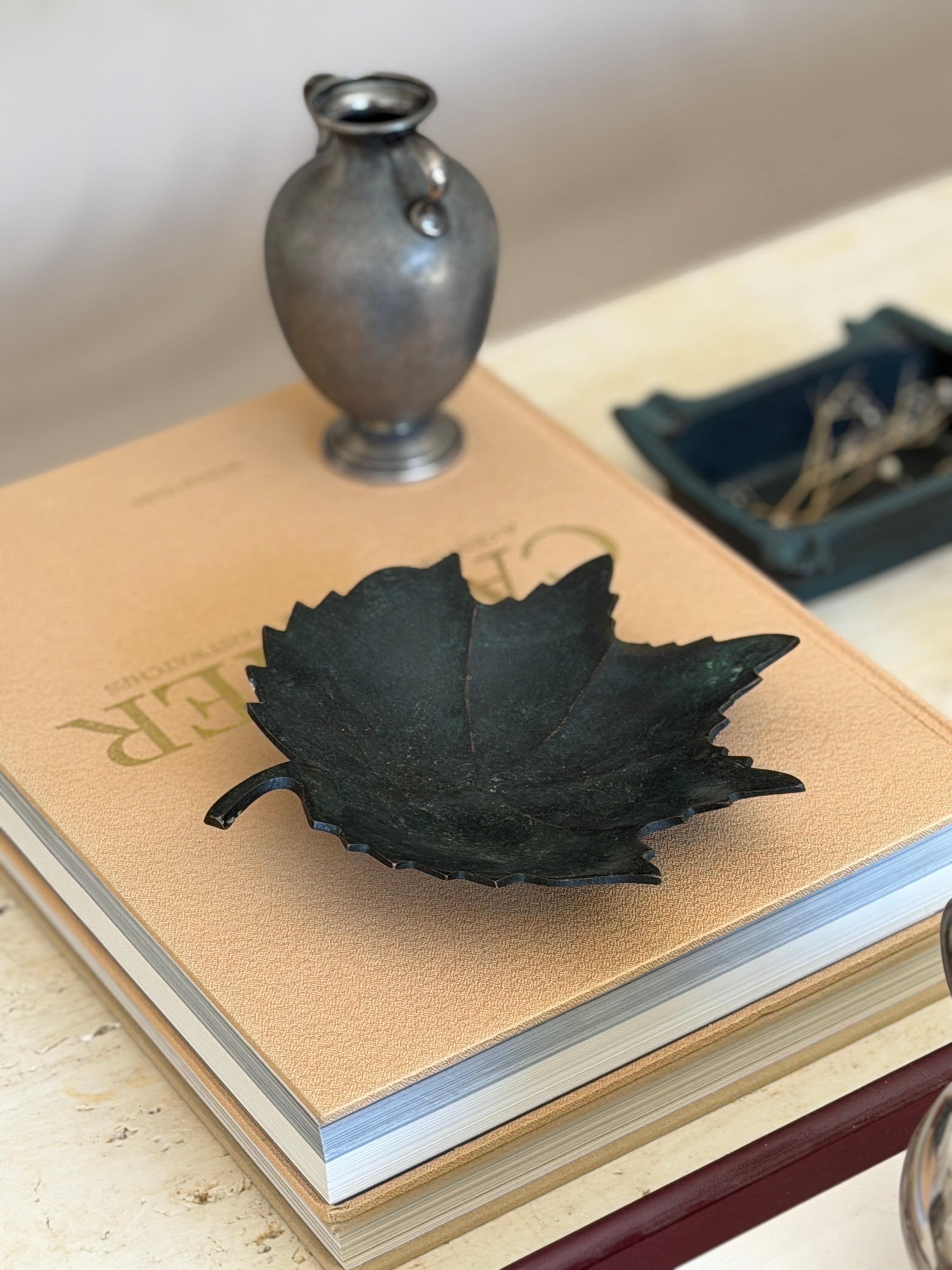 Angled desk display view of dark oxidized bronze leaf tray styled on a book stack alongside decor objects