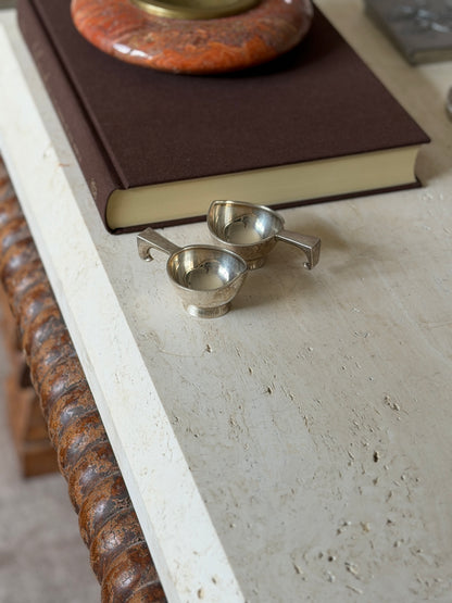 Two small silver dishes with angular geometric handles resting side by side on a stone surface, an open book above them