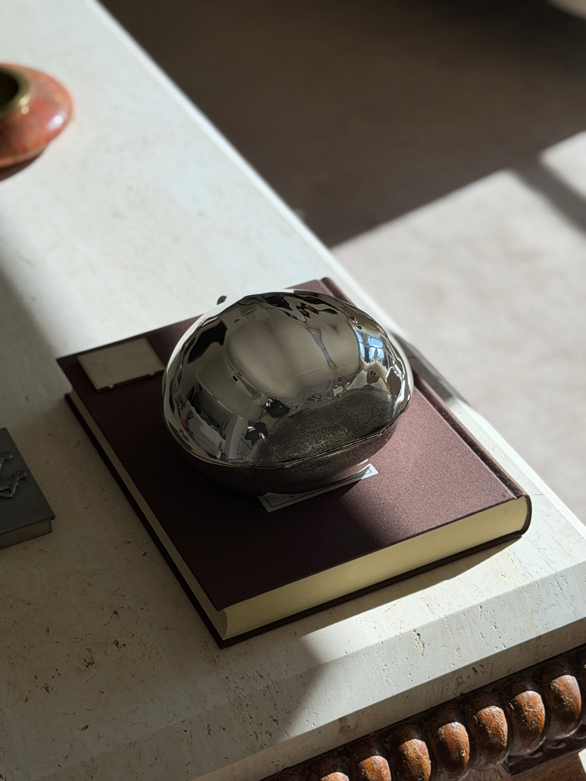 Overhead view of the Cartier sterling chestnut box on a brown book in raking sunlight, the textured shell tip at one side