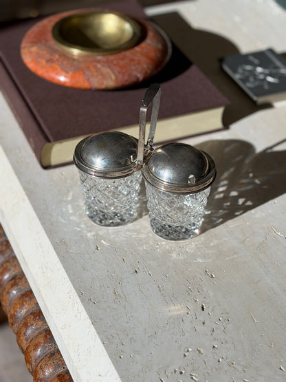 Cartier double jam pot with two diamond cut crystal jars under domed sterling lids joined by a tall center handle, on travertine in bright sun