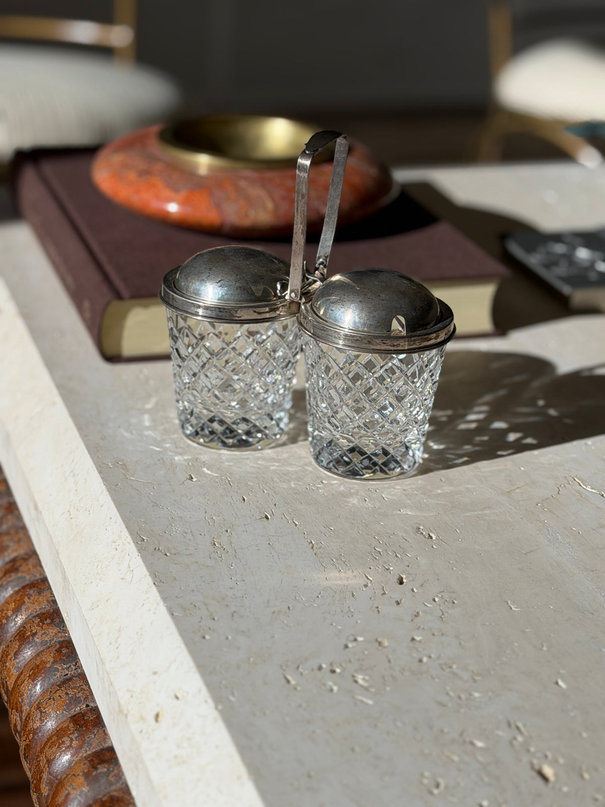 Front view of the twin crystal jam jars with hinged silver lids, one lid notched for a spoon, softly lit against a blurred brown book