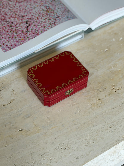 Closed red Cartier leather box with gold tooled border and clasp, resting on a stone surface beside an open book