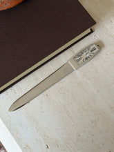 Full length view of the letter opener lying diagonally on a stone surface, its clock-face handle showing Roman numerals, with a book and stone bowl nearby