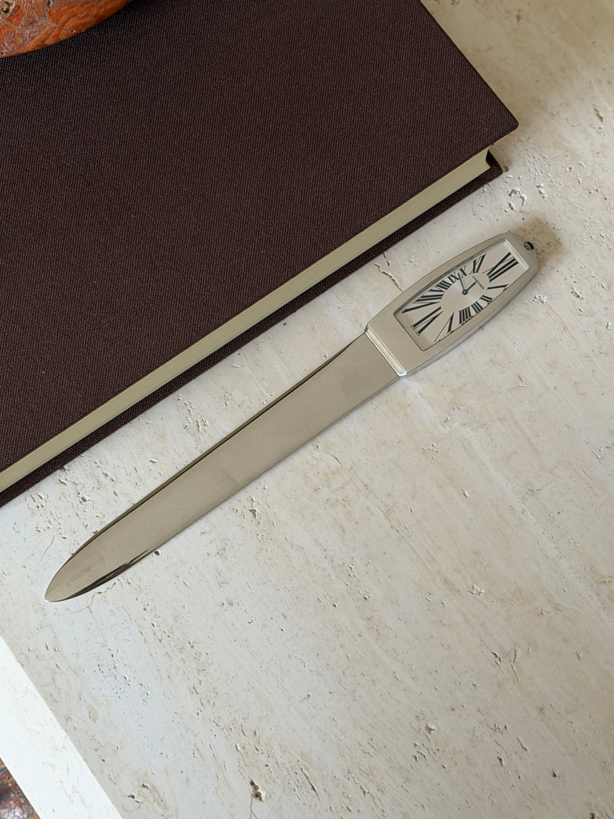 Full length view of the letter opener lying diagonally on a stone surface, its clock-face handle showing Roman numerals, with a book and stone bowl nearby