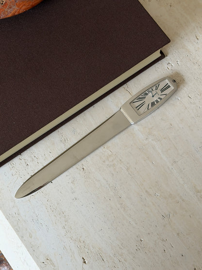 Full length view of the letter opener lying diagonally on a stone surface, its clock-face handle showing Roman numerals, with a book and stone bowl nearby