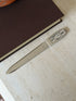 Full length view of the letter opener lying diagonally on a stone surface, its clock-face handle showing Roman numerals, with a book and stone bowl nearby