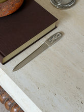 The letter opener shown from another angle on the stone surface beside the book, a woven silver-rimmed dish visible in the background