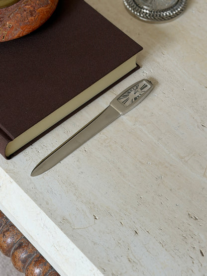 The letter opener shown from another angle on the stone surface beside the book, a woven silver-rimmed dish visible in the background