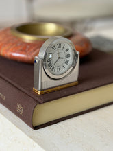 Cartier Ronde clock standing on a brown clothbound book, a red stone dish with brass insert softly blurred behind