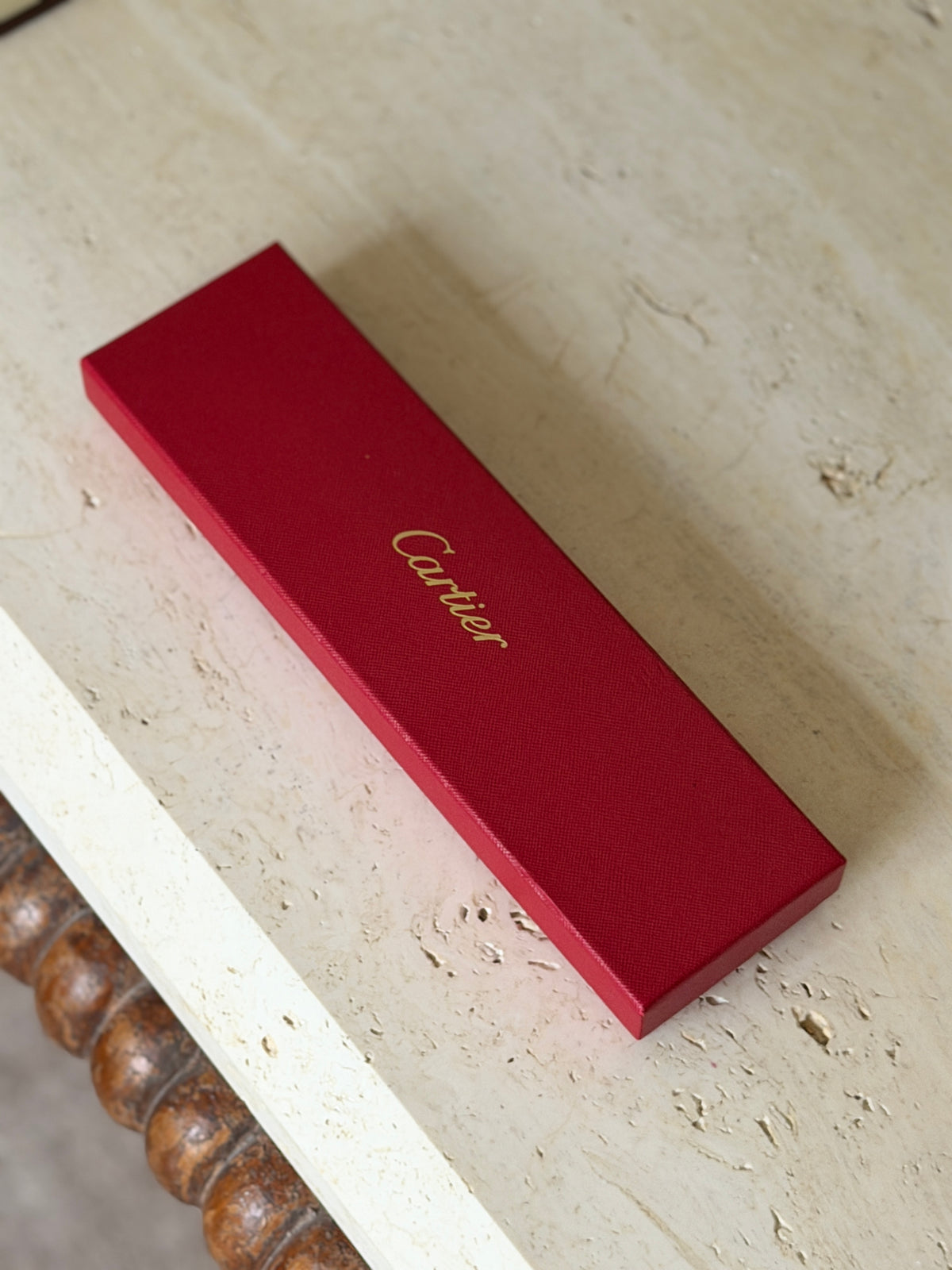 Original red Cartier presentation box with gold script logo on the grained lid, resting on a travertine table above a bobbin turned wood frame