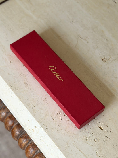 Original red Cartier presentation box with gold script logo on the grained lid, resting on a travertine table above a bobbin turned wood frame